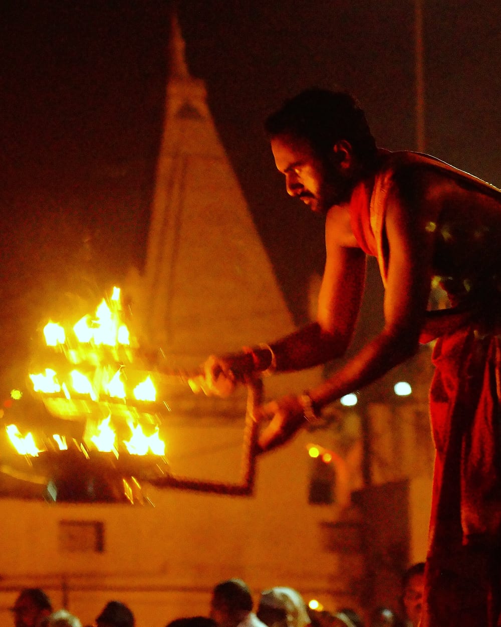 A tiered lamp of many flames held up in the dark at Gwarighat, the temple spire behind it.