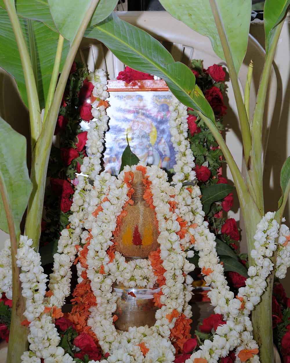 A pot topped with a coconut, wound in garlands of white and red flowers and framed by banana leaves.