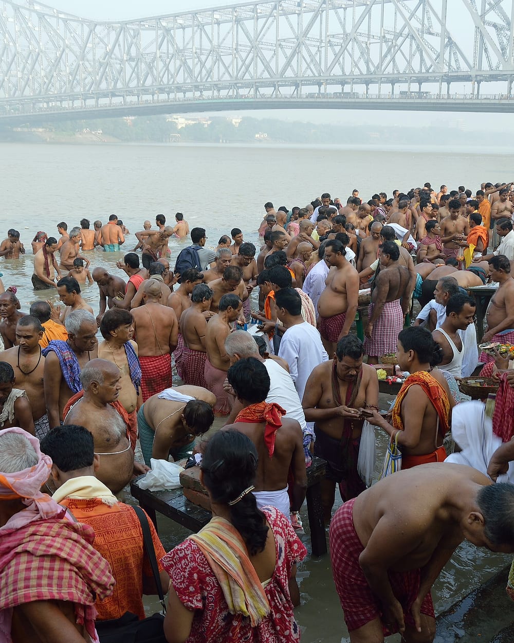 Men standing in the river at a Kolkata ghat, offering water to the ancestors, the Howrah Bridge behind.