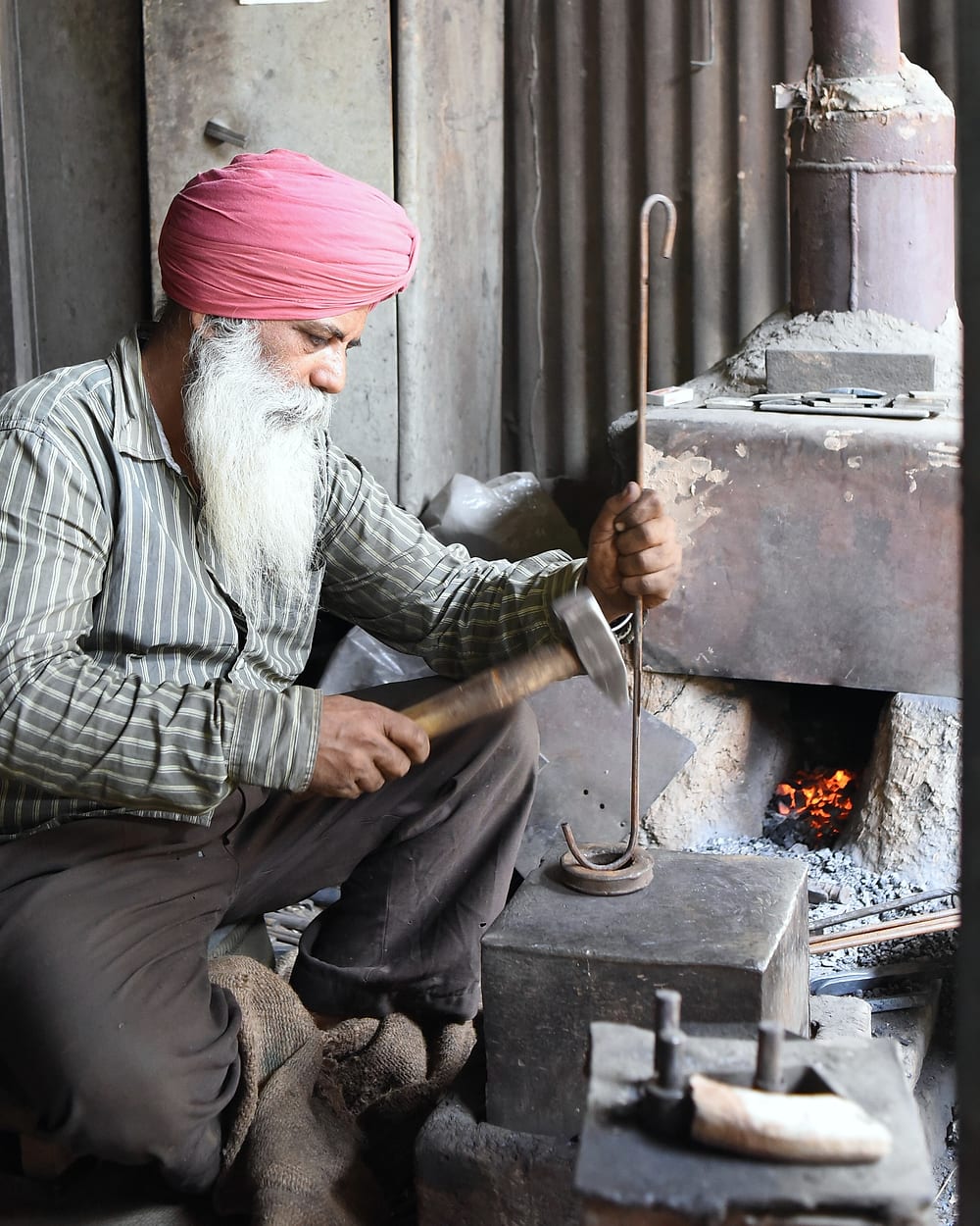 A bearded smith in a pink turban swings a hammer onto an iron rod at his forge.
