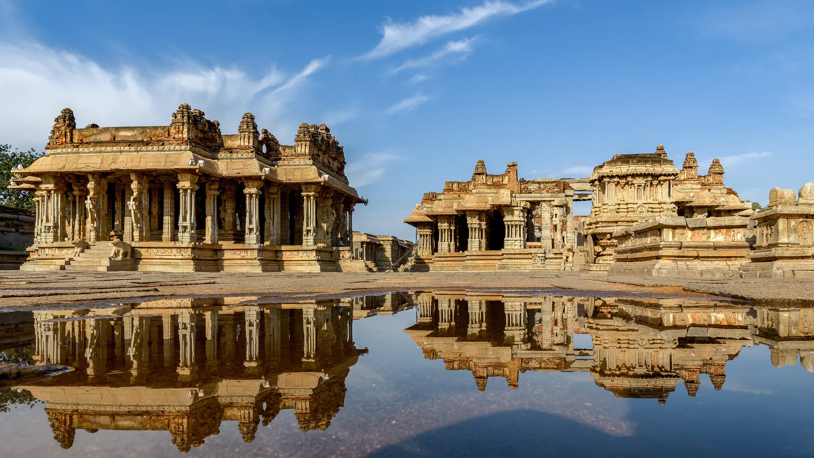 The stone pavilions of the Vitthala Temple at Hampi mirrored in a shallow pool under a blue sky