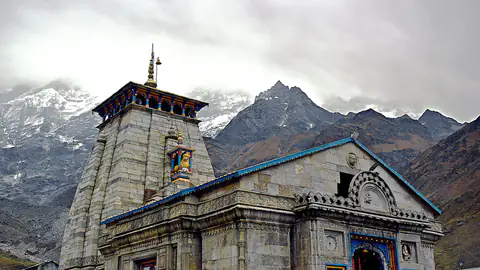 The grey stone Kedarnath temple with snow peaks behind it and pilgrims in the forecourt