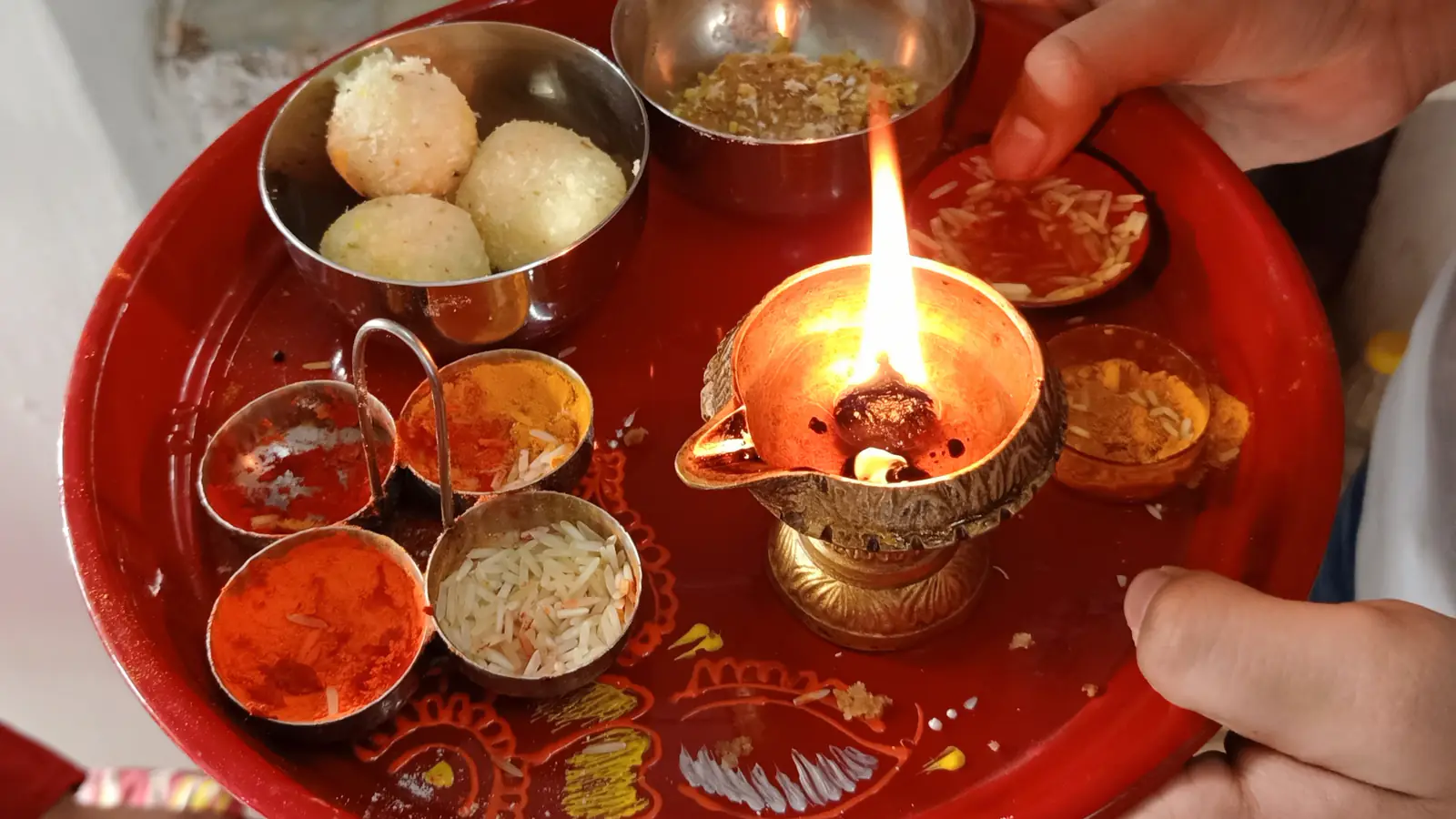 Hands hold a round red tray carrying a burning oil lamp, small bowls of kumkum and rice, flowers and sweets