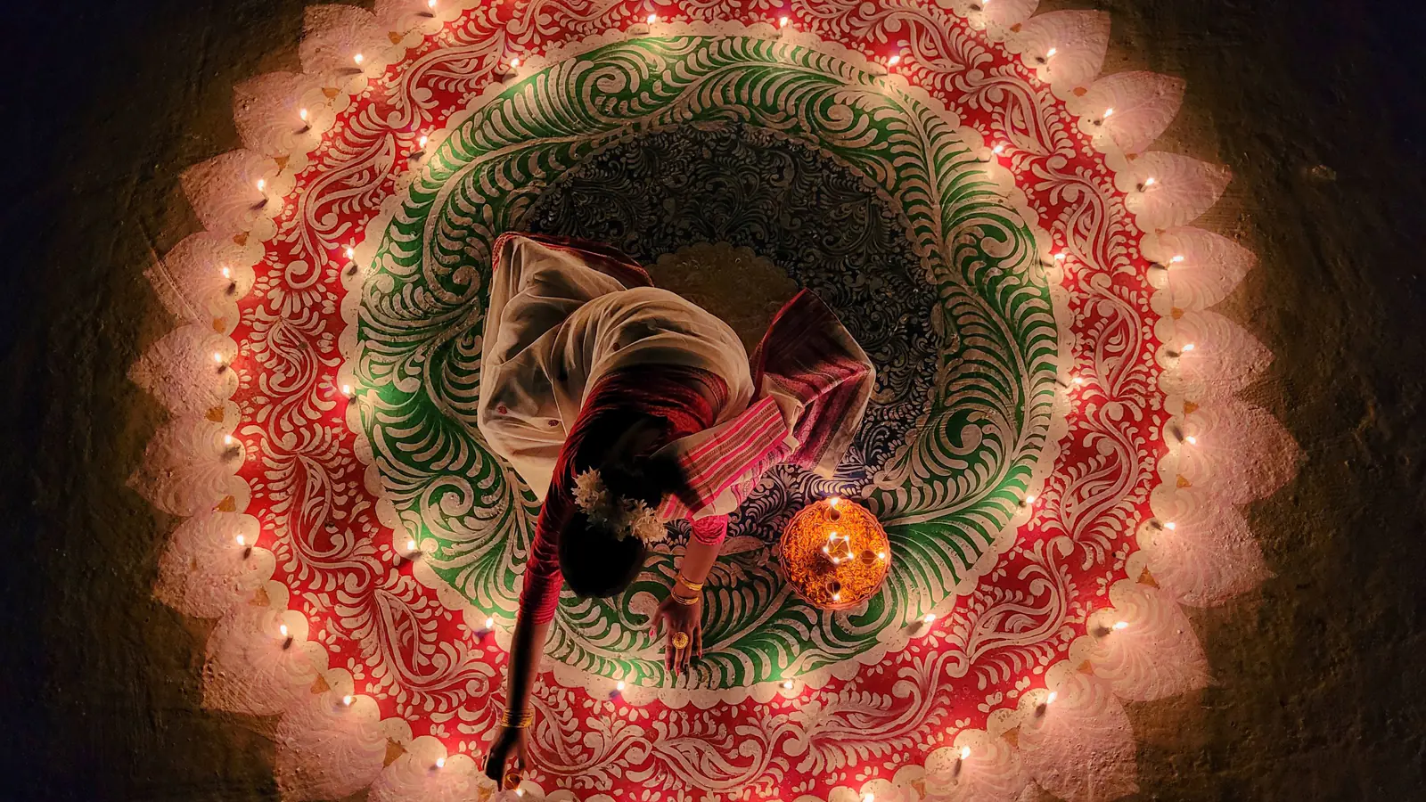 Seen from above, a woman lights a ring of small lamps set around a rangoli of leaves and petals