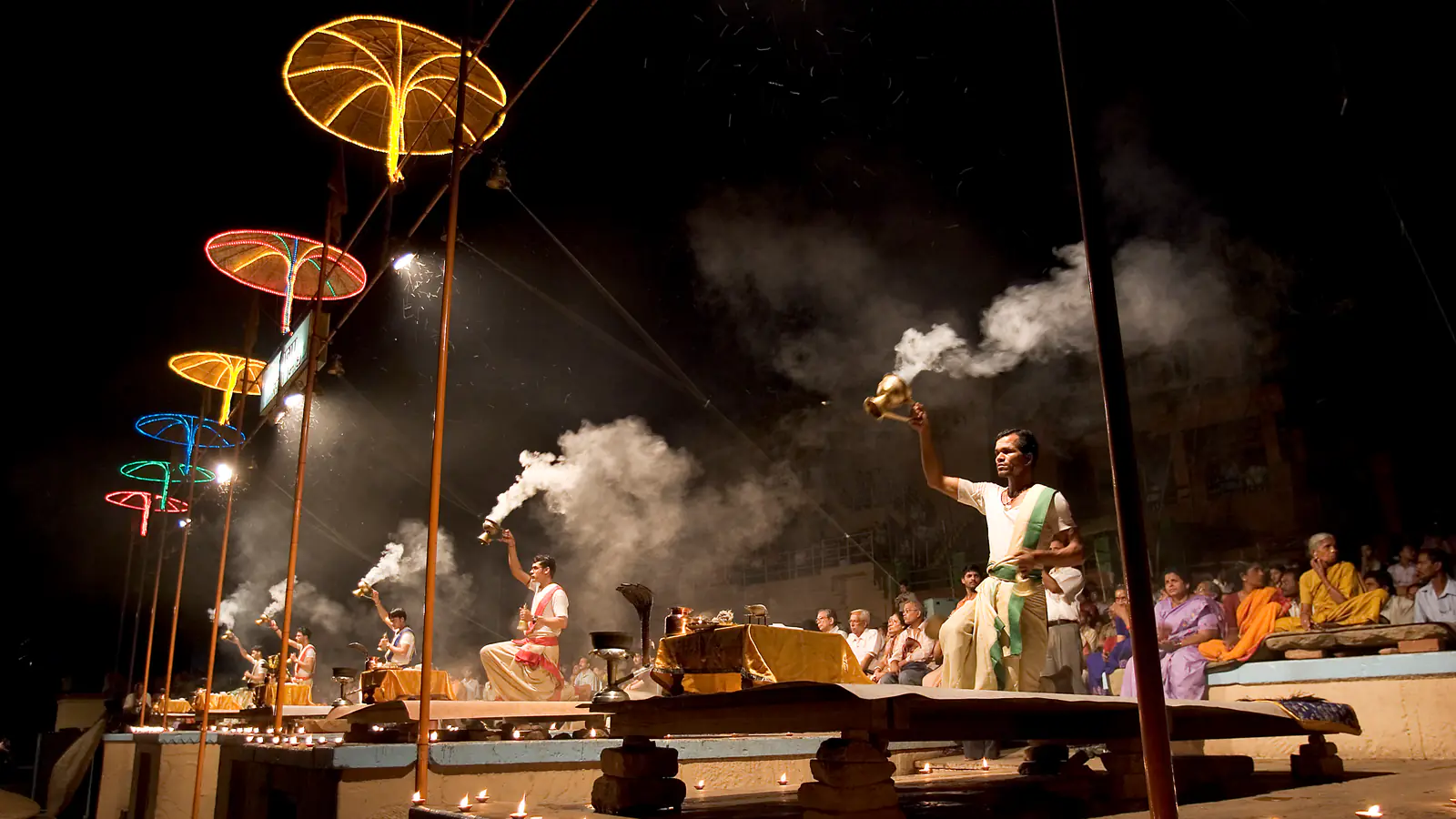 Priests raise smoking incense burners at night before a seated crowd, tall lamps lit above the platform