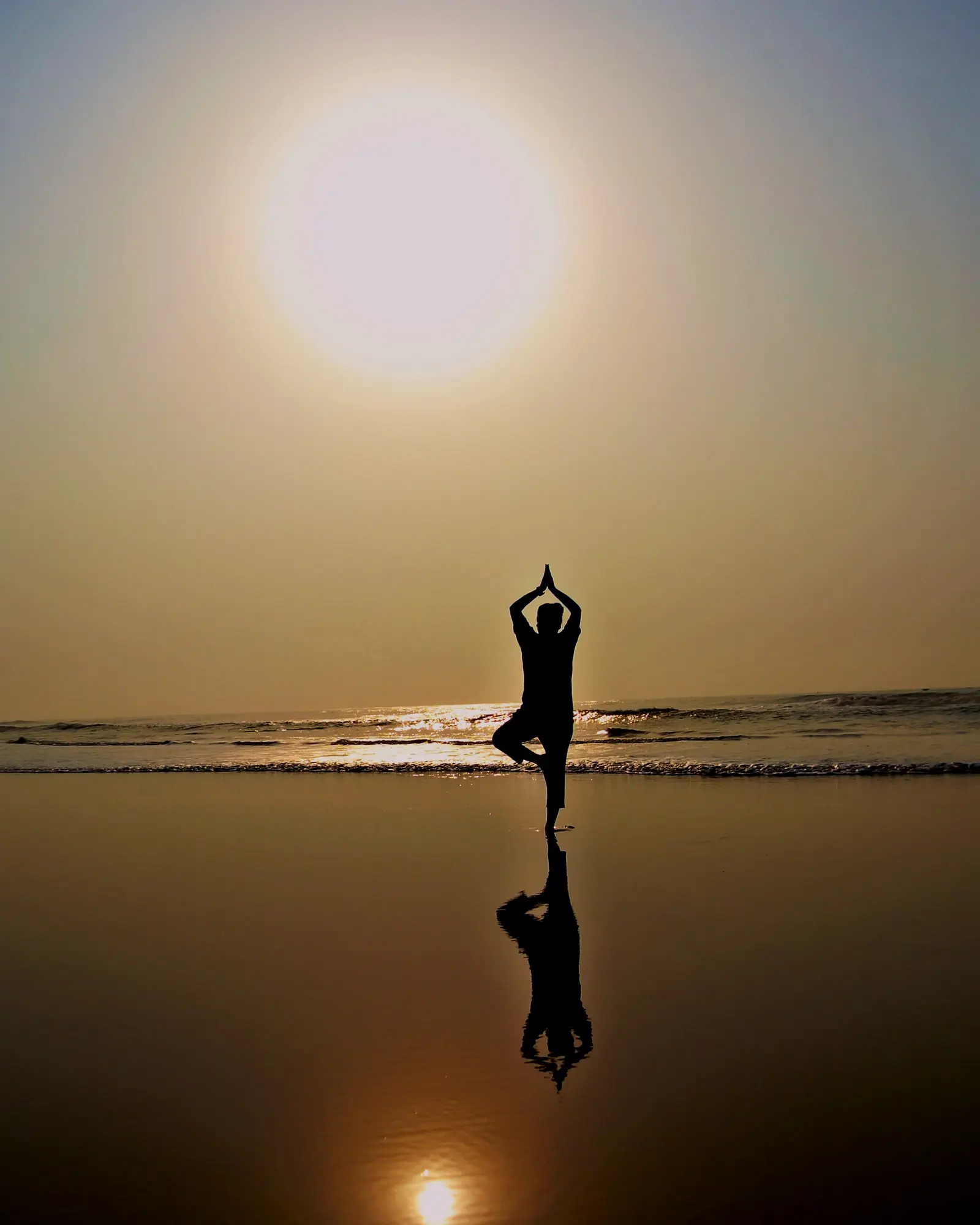 The silhouette of a person balancing in tree pose on a wet beach, the rising sun and its reflection behind